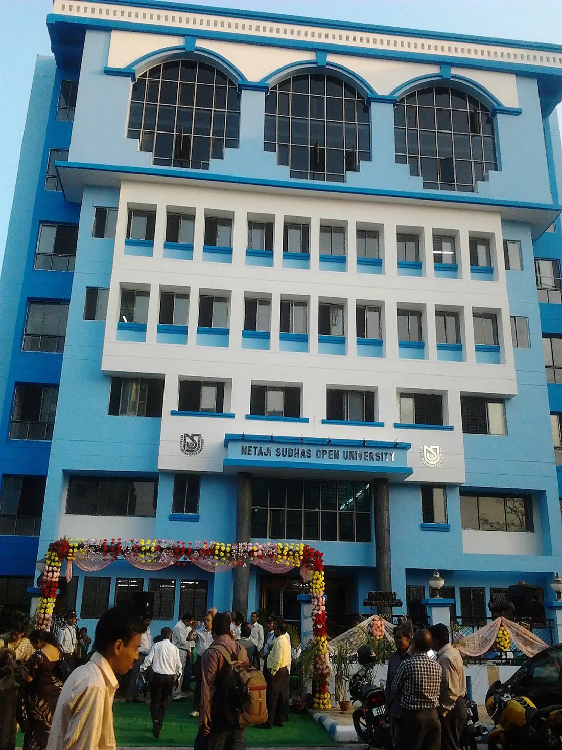 The blue and white headquarters building of NSOU, with people and a floral arch.